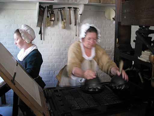 Two women in working womens 18th Century wear, working in a print shop. One is dabbing ink onto the type, which is sat in the printing bed or a wooden printing press, ready to print.